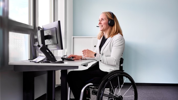 Die Parasportlerin Annika Zeyen-Giles sitzt in ihrem Rollstuhl im Büro an einem Tisch mit Bildschirm und Tastatur. Sie hat schulterlange blonde Haare, trägt einen weißen Blazer, eine schwarze Hose und schwarze Stiefeletten. Auf ihrem Kopf hat sie ein Headset, sie lächelt und befindet sich in einem Onlinemeeting.