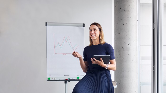 Die Paraleichtathletin Irmengard Bensusan sitzt in entspannt auf einem Hochstuhl vor einem Flipchart und hält ein I Pad in der Hand. Ihre schulterlangen Hellbraunen Haare sind offen, sie lächelt und trägt einen langen, dunkelblauen Rock und ein dunkelblaues T-Shirt.