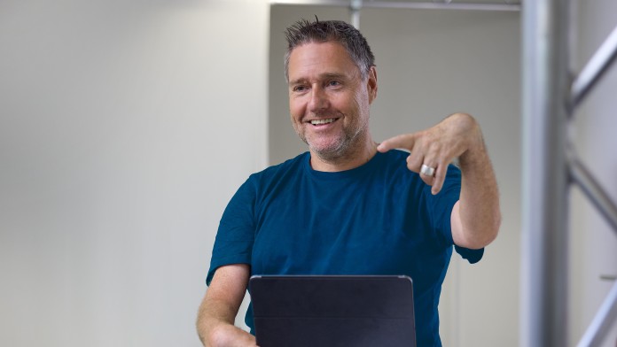 Der Para-Tischtennisspieler Jochen Wollmert steht an einem Tisch und hält ein IPad in der rechten Hand. Jochen Wollmert hat seit der Geburt eine Versteifung in den Hand- und Fußgelenken. Er trägt ein dunkelblaues T-Shirt und eine helle Jeans. Er lächelt freundlich in den Raum und hat kurze dunkelblonde Haare.