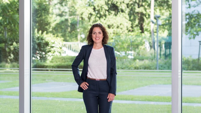 Marta Lambert, ist Teamleiterin in der IT. Sie trägt einen blauen Anzug mit einem weißen Hemd. Sie steht im Foyer des KfW Haupthaus Gebäudes in Frankfurt.