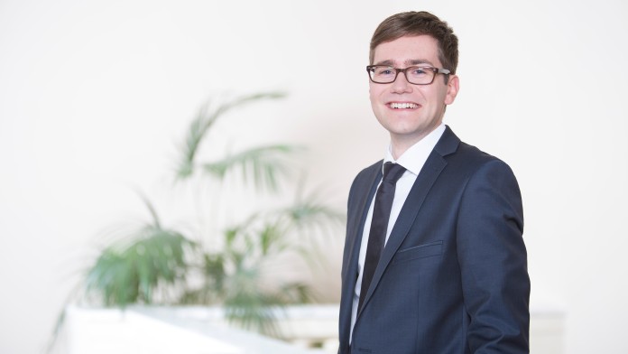 Benjamin Rother trägt einen blauen Anzug und eine Brille. Er befindet sich in dem historischen Berliner Marmor Saal des KfW Gebäudes in Berlin