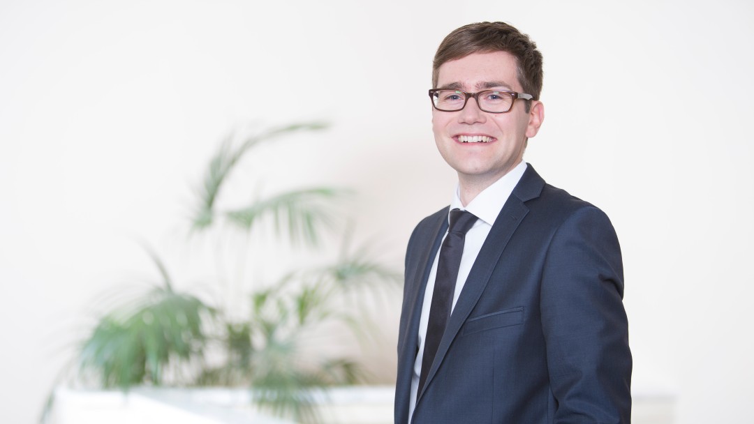 Benjamin Rother trägt einen blauen Anzug und eine Brille. Er befindet sich in dem historischen Berliner Marmor Saal des KfW Gebäudes in Berlin