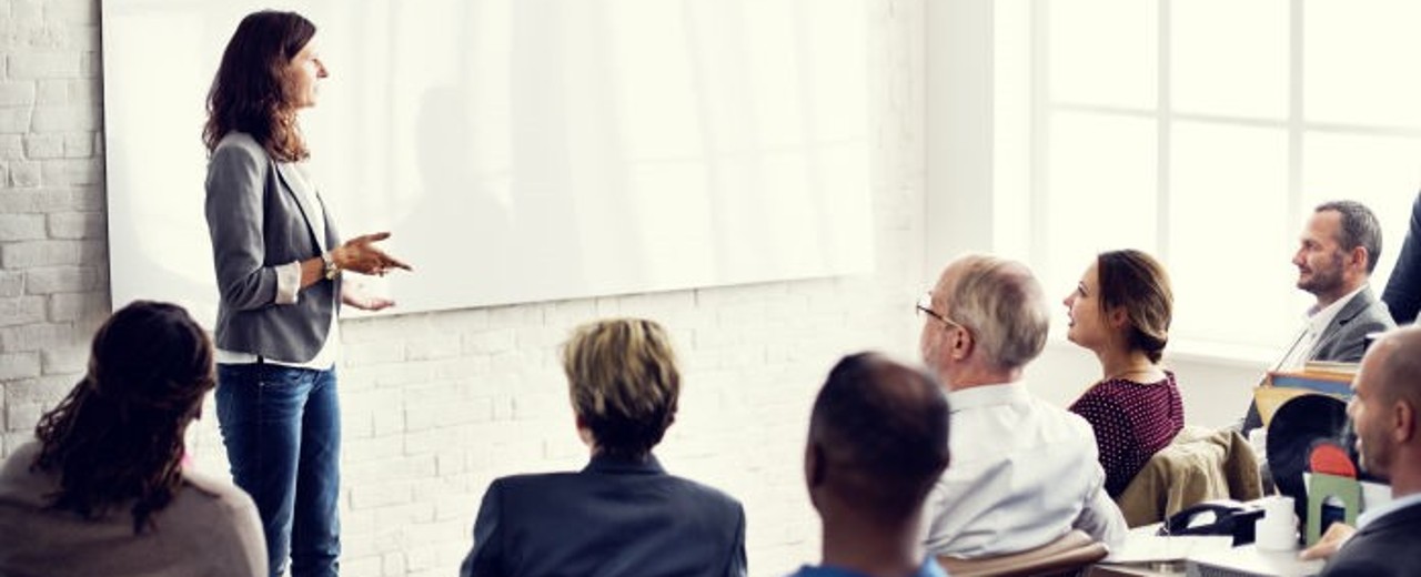 Mehrere Mitarbeitenden nehmen an einer Schulung bzw. Workshop teil. Eine Frau moderiert den Workshop und steht vor den sitzenden ihr aufmerksam zugewandten Teilnehmenden.