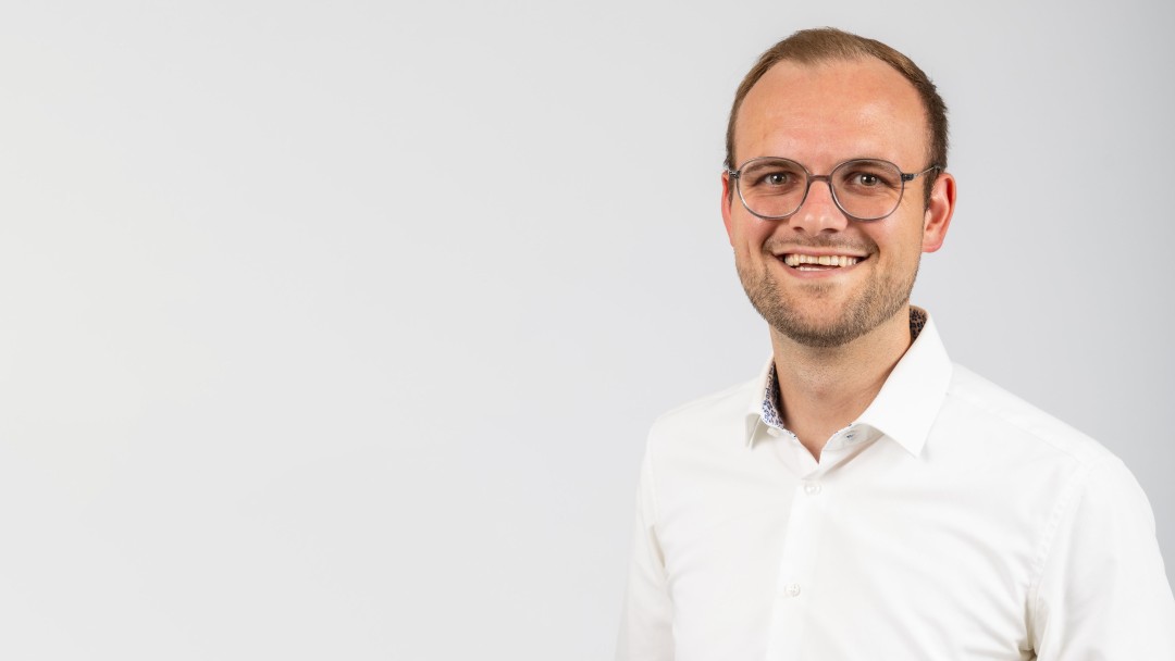 Lukas Muders, Mitarbeiter der KfW Bankengruppe, steht lächelnd vor einem weißen Hintergrund. Er trägt ein weißes Hemd und eine Brille. 