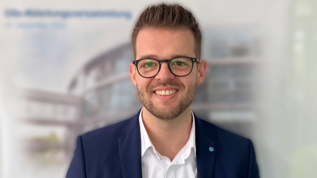 Marc Hammer, ein Mitarbeiter der KfW Bankengruppe am Standort Bonn, lächelt indie Kamera. Er trägt eine Brille, ein weißes Hemd und einen blauen Sakko.