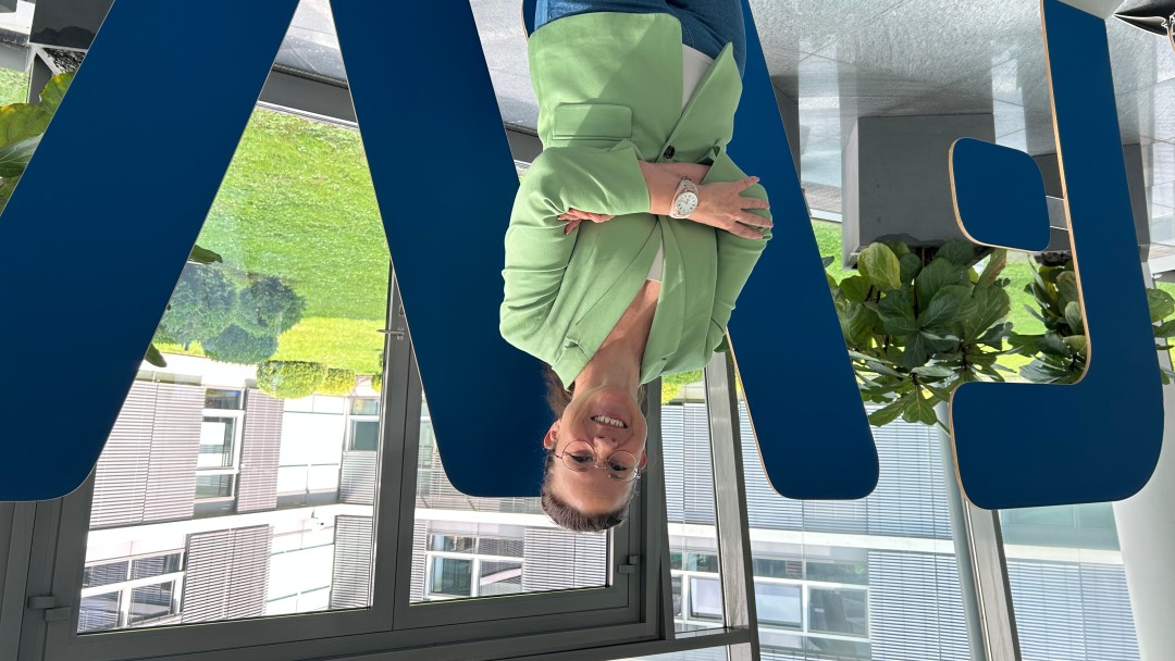 Nicole Banse, Mitarbeiterin der KfW Bankegruppe am Standort Bonn, steht im Foyer mit verschränkten Armen und lächelt. Sie trägt eine Brille und einen grünen Blazer. Im Hintergrund ist der begrünte Innenhof zu sehen. 