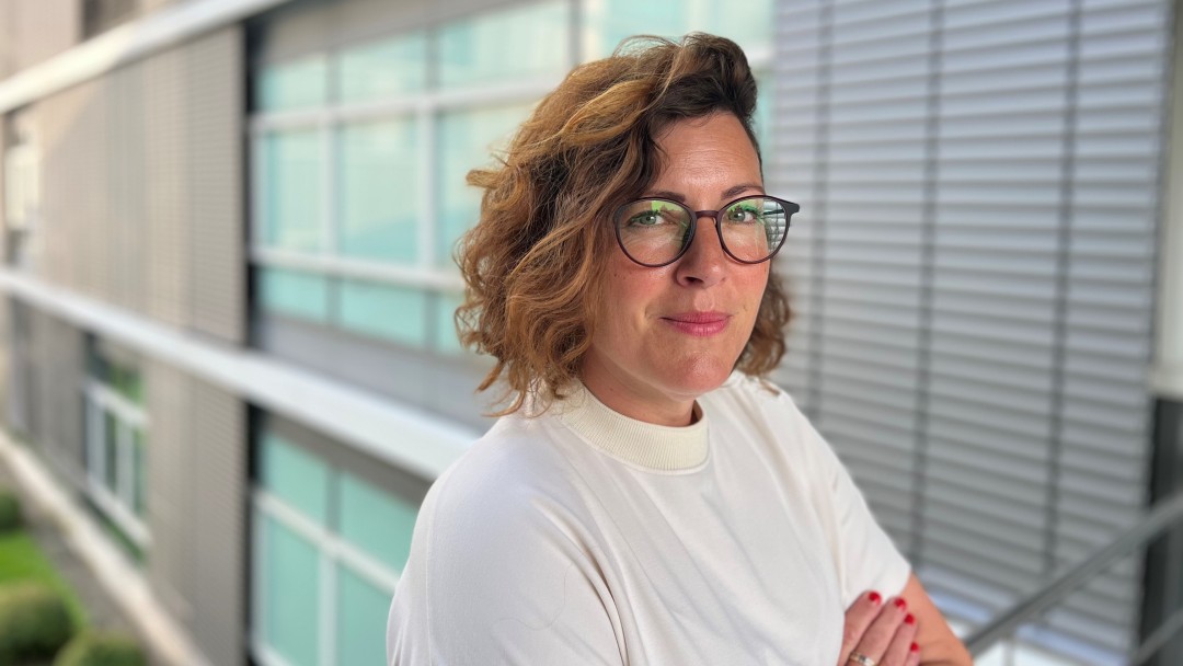 Veronika Diekmann, Mitarbeiterin der KfW Bankengruppe am Standort Bonn, steht mit verschränkten Armen im Freien vor einem Gebäude im Grünen. Sie trägt ein helles T-Shirt, kinnlange Haare und eine Brille, und schaut in die Kamera.