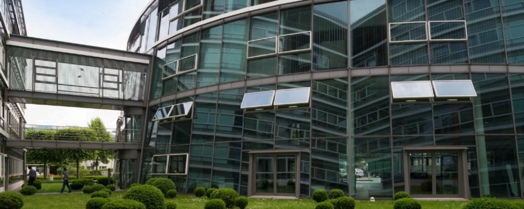 Empfangsbereich der KfW-Niederlassung in Bonn in einem großen verglasten Foyer mit grauen Fliesen, farbigen Balken in Weiß, Blau und Orange. Zwei Personen sitzen am Empfang. Weitere Menschen bewegen sich durch den Raum, im Hintergrund Grünfläche.