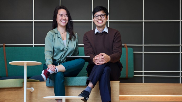 Fabian Nguyen und eine Kollegin, Mitarbeitende der KfW Bankengruppe und Mitglieder des internen Asian-Netzwerk, sitzen mit überschlagenen Beinen in der Green Lounge der KfW Westarkade und lächeln in die Kamera. 