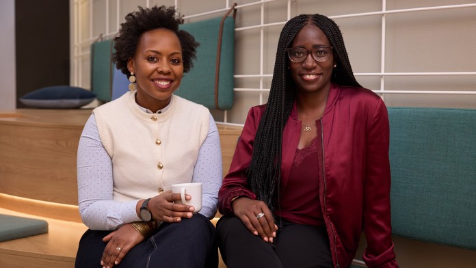 Zwei Kolleginnen der KfW Bankengruppe und Mitglieder des internen People-of-Color-Netzwerk, sitzen mit überschlagenen Beinen in der Green Lounge der KfW Westarkade und lächeln in die Kamera. 