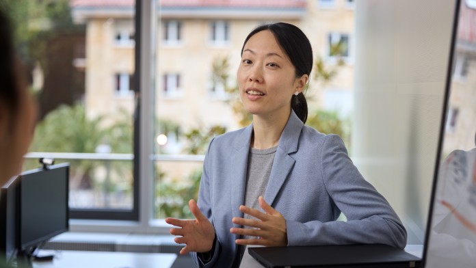 Lea Cho, Mitarbeiterin der KfW Bankengruppe, steht hinter einem Pult mit einem Laptop und ist im Gespräch mit einer Person außerhalb des Bildausschnitts. Sie trägt einen hellblauen Blazer und ein graues Shirt. 