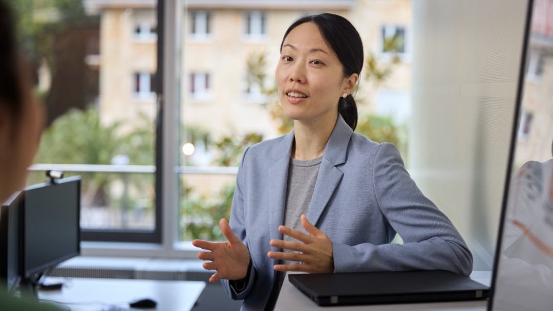 Lea Cho, Mitarbeiterin der KfW Bankengruppe, steht hinter einem Pult mit einem Laptop und ist im Gespräch mit einer Person außerhalb des Bildausschnitts. Sie trägt einen hellblauen Blazer und ein graues Shirt. 