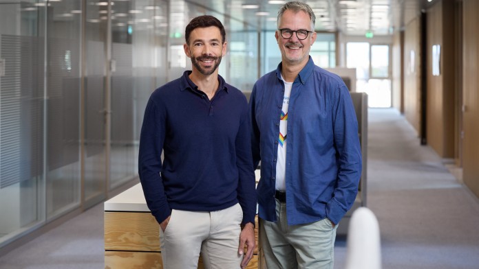 Michael Kemper und Christoph Seibert, Mitarbeitende der KfW Bankengruppe und Mitglieder des internen Rainbow-Netzwerkes, stehen im Flur vor einem hohen Tischpult und lächeln in die Kamera.