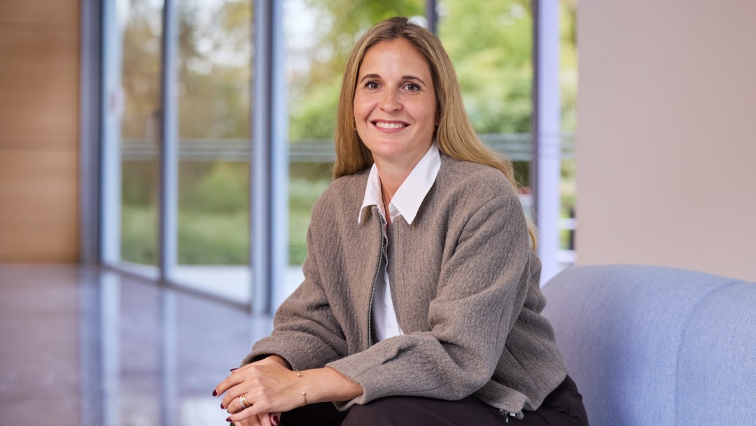 Sandra Freudenberg, Mitarbeiterin der KfW Bankengruppe, sitzt auf einem hellblauen Sofa. Sie trägt eine dunkelblaue Hose, eine hellbraune Jacke und ein weißes Hemd. Sie hat dunkelblonde lange Haare und lächelt in die Kamera. 