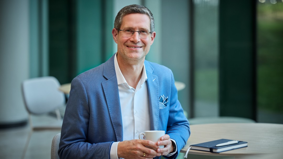 Thomas Kirsch, Mitarbeiter der KfW Bankengruppe, sitzt an einem Tisch und hält eine weiße Tasse in der Hand. Er trägt einen blauen Sakko, ein weißes Hemd und eine Brille und lächelt in die Kamera. Auf dem Tisch liegt ein Notizbuch mit einem Kugelschreiber. 