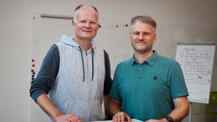 Zwei Mitarbeiter der KfW Bankengruppe und Mitglieder des internen Väternetzwerks stehen hinter einem hohen Tischpult und lächeln in die Kamera. Auf dem Tisch liegen zwei Notizbücher.
