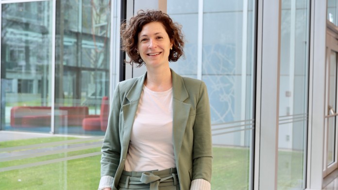 Portraitbild von Alena von Rüden, Mitarbeiterin der Internen Revision der KfW, sie trägt einen olivgrünen Blazer mit farblich passender Hose und steht in einem Foyer der KfW.