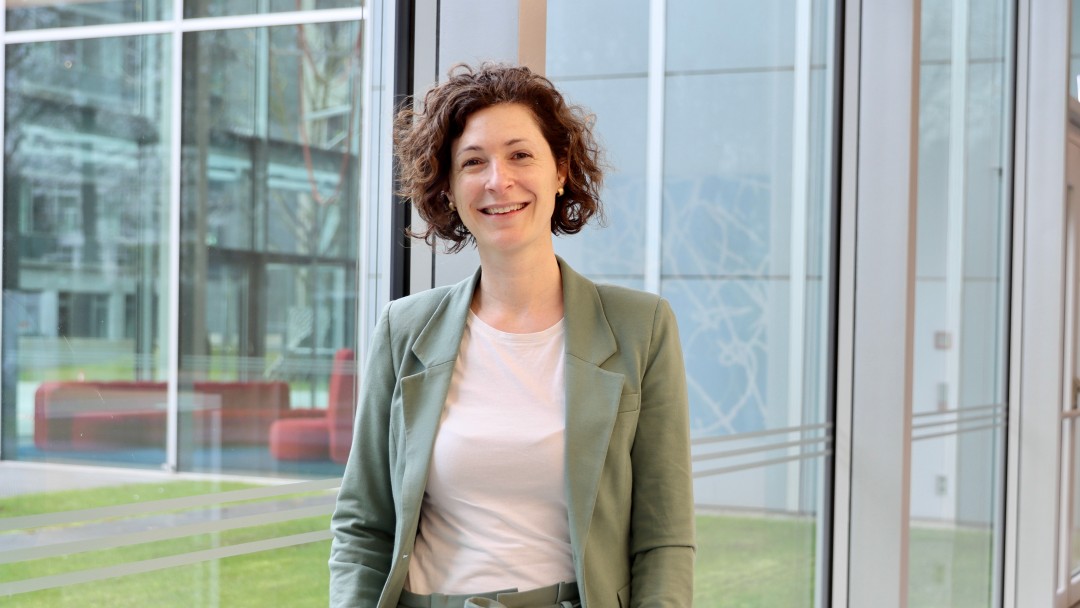 Portraitbild von Alena von Rüden, Mitarbeiterin der Internen Revision der KfW, sie trägt einen olivgrünen Blazer mit farblich passender Hose und steht in einem Foyer der KfW.