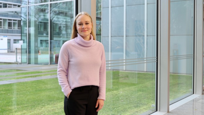 Portraitbild von Alexandra Dziemba, Mitarbeiterin der Internen Revision der KfW, sie trägt einen hellrosanen Rollkragenpullover sowie dunkle Hose und steht in einem Foyer der KfW.