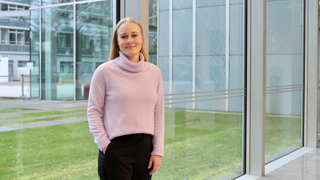 Portraitbild von Alexandra Dziemba, Mitarbeiterin der Internen Revision der KfW, sie trägt einen hellrosanen Rollkragenpullover sowie dunkle Hose und steht in einem Foyer der KfW.