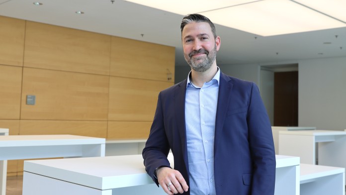 Portraitbild von Christian Heizmann, Mitarbeiter der Internen Revision der KfW, er trägt ein hellblaues Hemd mit dunkelblauem Sakko und steht in einem Foyer der KfW.