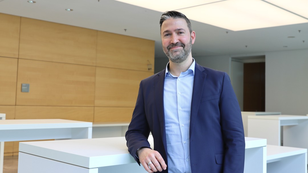 Portraitbild von Christian Heizmann, Mitarbeiter der Internen Revision der KfW, er trägt ein hellblaues Hemd mit dunkelblauem Sakko und steht in einem Foyer der KfW.