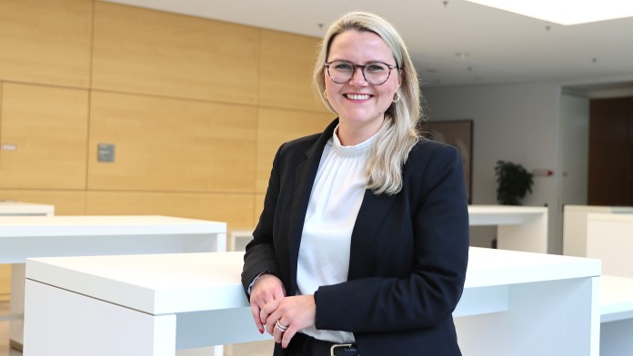 Portraitbild von Jana Alina Polzin, Mitarbeiterin der Internen Revision der KfW, sie trägt eine Brille sowie ein weißes Oberteil mit dunklem Blazer und steht in einem Foyer der KfW.