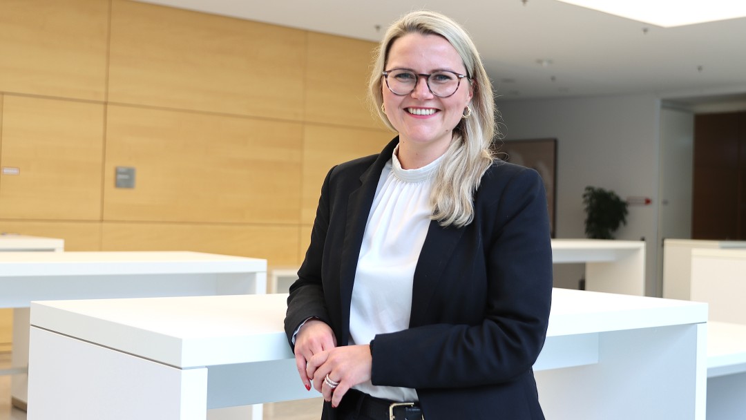 Portraitbild von Jana Alina Polzin, Mitarbeiterin der Internen Revision der KfW, sie trägt eine Brille sowie ein weißes Oberteil mit dunklem Blazer und steht in einem Foyer der KfW.