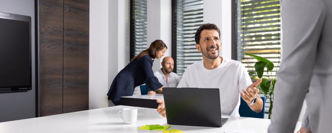Ein Mann sitzt an einem Tisch und unterhält sich mit einer Frau, die vor ihm steht und sich mit den Händen auf dem Tisch abstützt. Auf dem Tisch steht ein Laptop und eine Kaffeetasse. Außerdem liegen dort ein paar Post-Its und ein Block. Im Hintergrund sind zwei weitere Personen zu sehen, die sich miteinander unterhalten. 