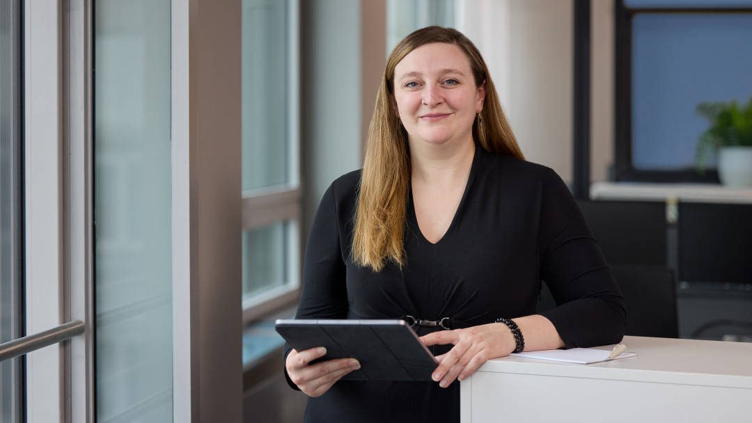 Chelsea Ashby, Mitarbeiterin der KfW Bankengruppe, steht angelehnt an einem weißen Pult, hält ein iPad in der Hand und lächelt in die Kamera. Sie trägt ein schwarzes Outfit und hat lange helle Haare.