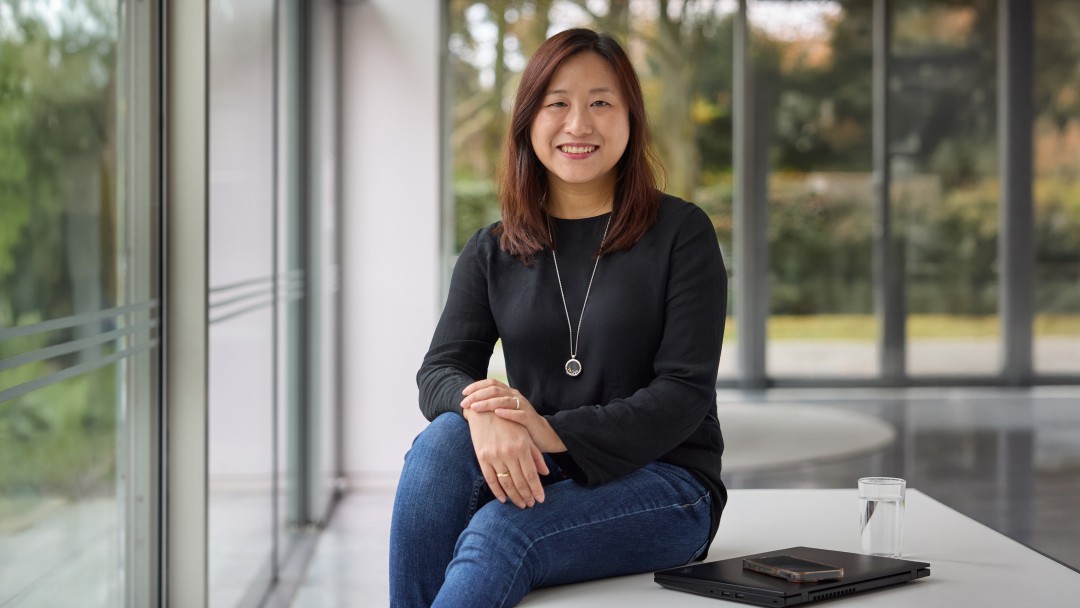 Hoang Hong Ngoc, Mitarbeiterin der KfW Bankengruppe, sitzt lächelnd auf einer weißen Flächevor einer Fensterfront mit Blick ins Grüne. Neben liegt ein zugeklappter Laptop, ein Handy und ein Wasserglas. Sie trägt eine blaue Jeans und ein schwarzes Oberteil. Sie hat schulterlange braune Haare. 