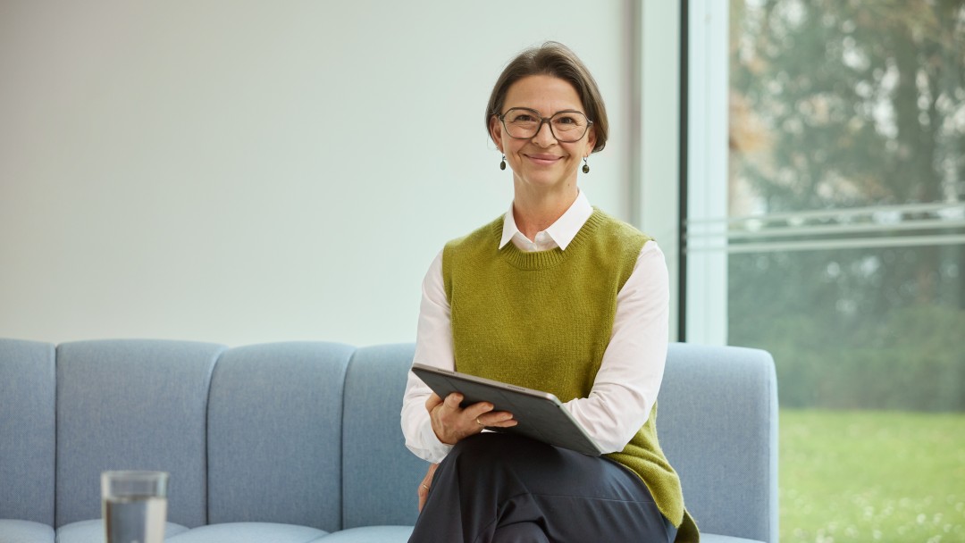 Isabelle Bobzin, Mitarbeiterin der KfW Bankengruppe, sitzt auf einem hellblauem Sofa vor einer großen Fensterfront mit Gartenblick. Vor ihr steht ein schwarzer runder Tisch mit Handy und einem vollen Glas Wasser. Sie hält ein iPad , trägt eine dunkle Hose, ein weißes Hemd und einen grünen Pullunder. Sie hat kurze Haare, trägt eine Brille und lächelt in die Kamera. 