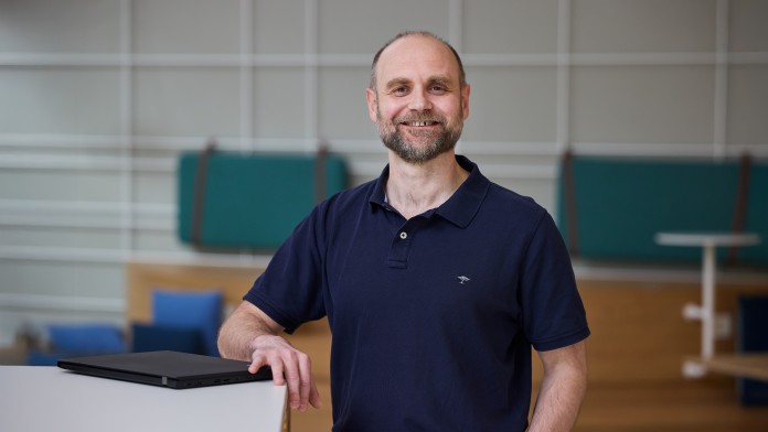 Jens Schöbel, Mitarbeiter der KfW Bankengruppe, steht an einem Hochtisch und lächelt freundlich in die Kamera. Auf dem Tisch ist ein Laptop. Im Hintergrund sind Sitzmöglichkeiten. 