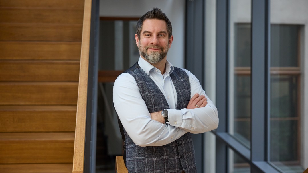 Jeroen Schäffer, Mitarbeiter der KfW Bankengruppe, steht mit verschränkten Armen neben einer Holztreppe und blickt in die Kamera. Er trägt ein helles Hemd mit grauer karierter Weste, eine Armbanduhr und hat kurze dunkele Haare sowie einen helleren Vollbart. 