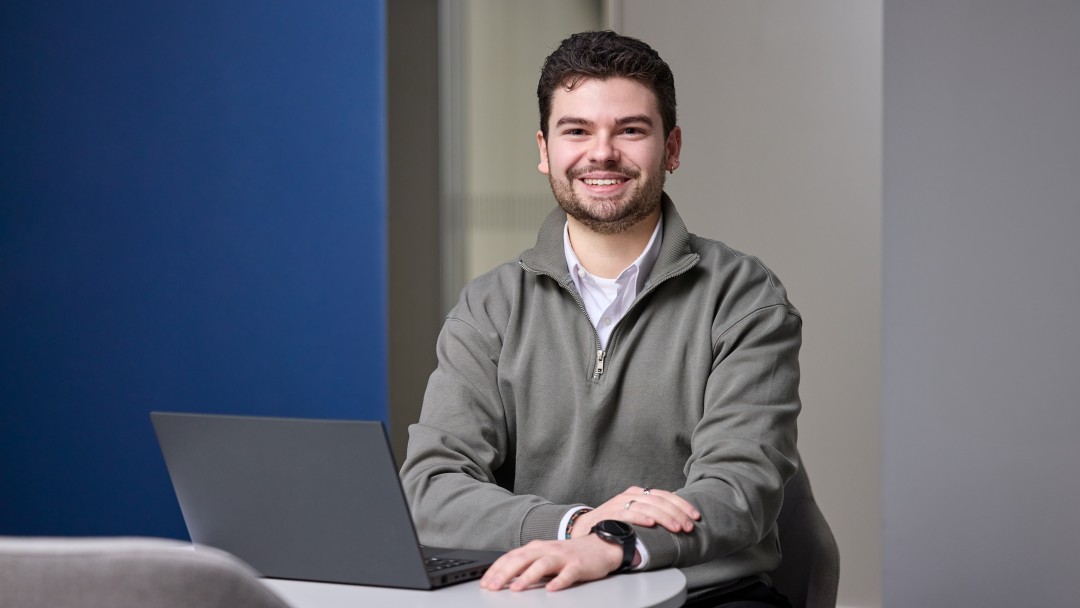 Pio Guiseppe Barberi, Mitarbeiter der KfW Bankengruppe, sitzt lächenld an einem hohen weißen Tischvor einem blau-weißen Hintergrund. auf dem Tisch steht ein augeklappter schwarzer Laptop. er trägt ein weißes Hemd mit einem dunklen Sweater darüber. Er hat kurze braune Haare und trägt eine schwarze Armbanduhr.