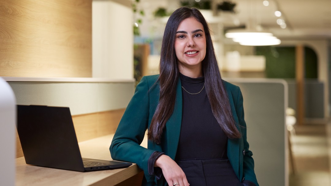 Sabrina Naimi, Mitarbeiterin der KfW Bankengruppe, steht an einem hohem Pult mit einem aufgeklappten Laptop. Sie trägt ein schwarzes Outfit und dazu einen dunkelgrünen Blazer. Sie hat langebraune Haare und lächelt in die Kamera.