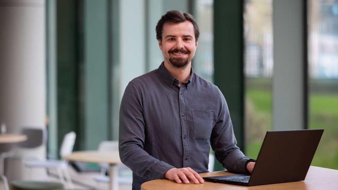Wilhelm Teicke, Mitarbeiter der KfW Bankengruppe, steht an einem Hochtisch mit aufgeklapptem Laptop und lächelt in die Kamera. Im Hintergrund ist eine Fensterfront mit Blick ins Grüne.