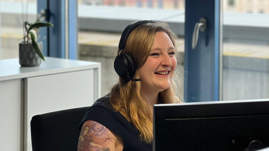 Chelsea Ashby, Mitarbeiterin der KfW Bankengruppe, sitzt freundlich lächelnd an ihrem Arbeitsplatz mit schwarzem T-Shirt und Headset vor dem Monitor. Im Hintergrund sind ein Sideboard, eine Fensterfront und eine kleine Pflanze zu sehen. 
