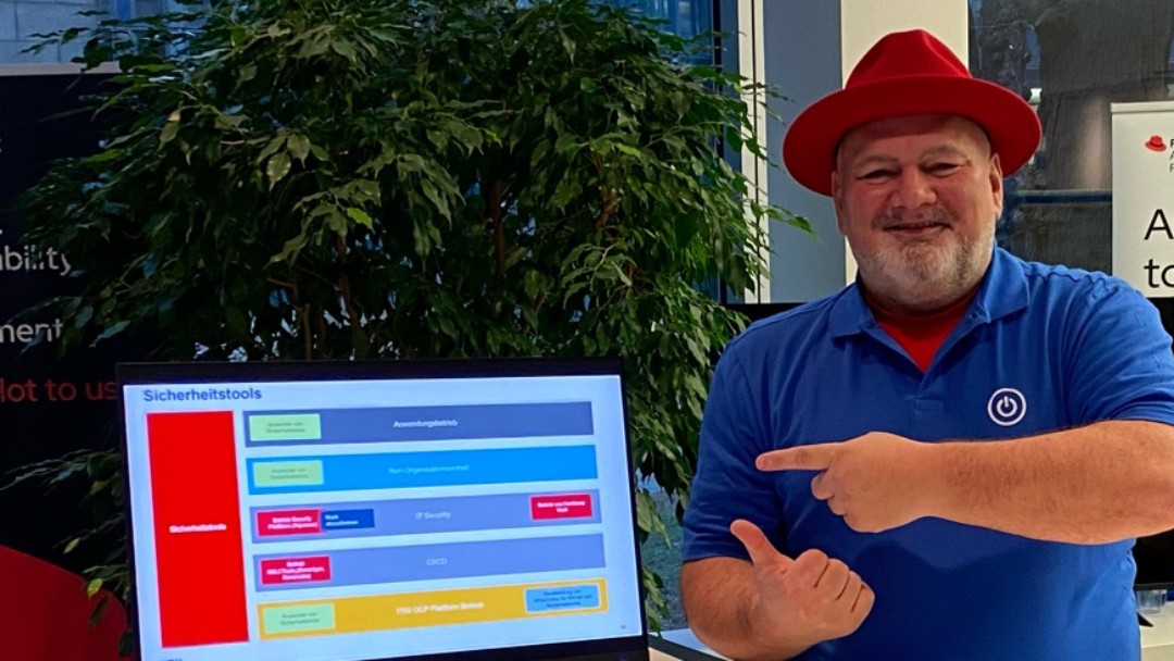 Heino Jesuiter, Mitarbeiter der KfW Bankengruppe, befindet sich an einem Stand einer KfW internen Messe zum Thema IT-Infrastruktur, er trägt ein blaues Polo-Shirt und einen roten Hut.