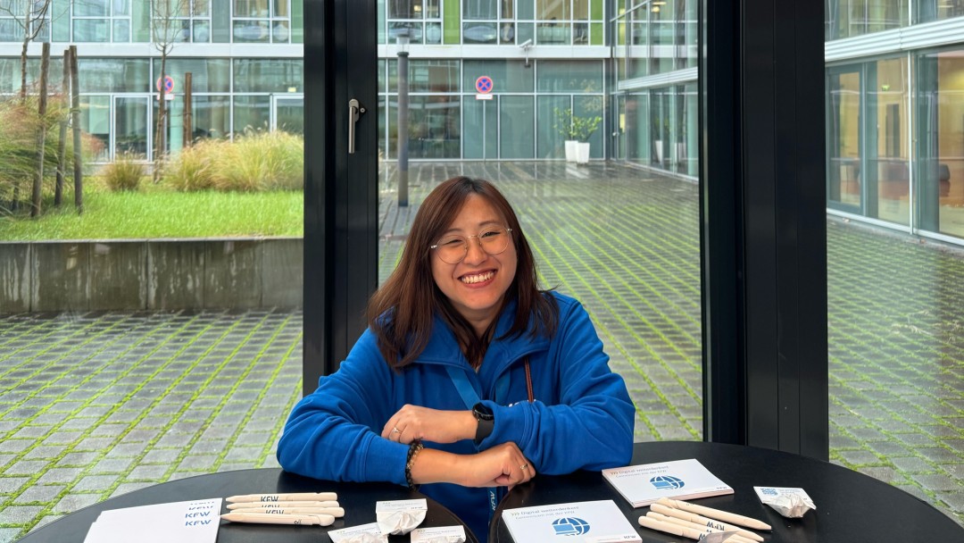 Hong Ngoc Hoang, Mitarbeiterin der KfW Bankengruppe, steht lächelnd hinter zwei schwarzen Stehtischen mit KfW-Werbeartikeln. Sie trägt einen blauen Pullover, hat schulterlange braune Haare und eine Brille. Im Hintergrund ist eine Fensterfront mit Blick auf den begrünten Innenhof der KfW zu sehen. 