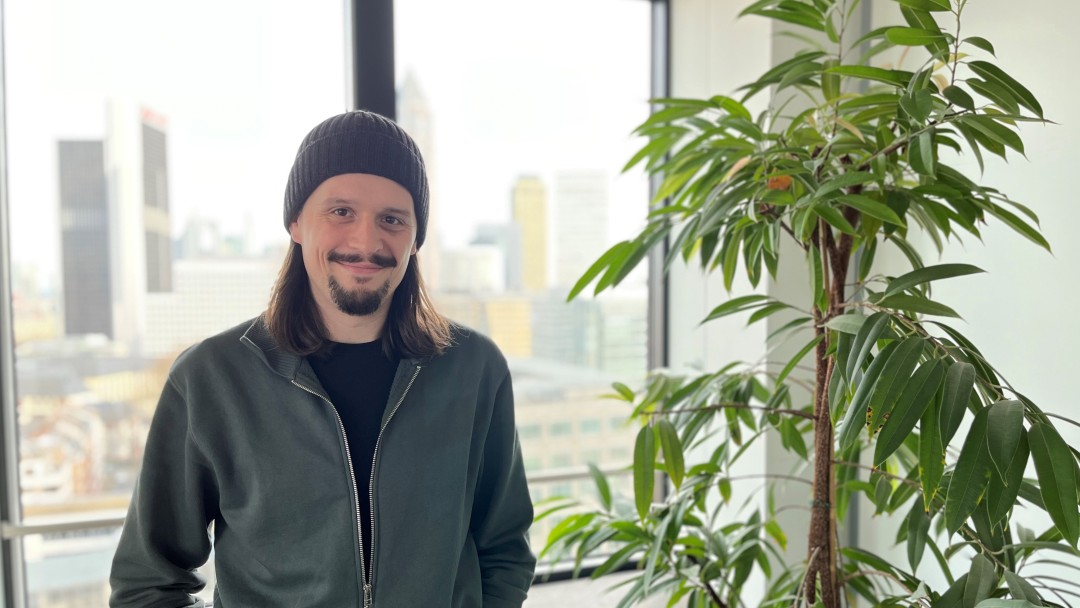 Portraitbild von Philipp Ganz, Mitarbeiter aus dem IT-Bereich KfW. Er hat dunkle kinnlange Haare und einen Bart, trägt eine Mütze und grauen Pullover mit Reißverschluss.