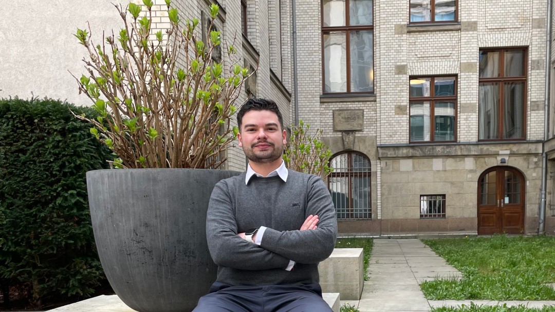 Ein Mitarbeiter der KfW Bankengruppe, Pio Giuseppe Barberi, sitzt draußen auf einer Mauer, imHintergrund sind ein Gebäude und eine grüne umgebung zu sehen. 