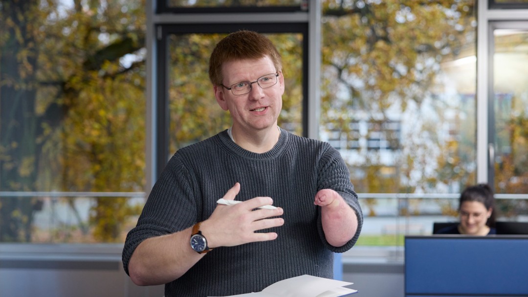 Daniel Metz, Mitarbeiter der KfW Bankengruppe, trägt einen grauen Pullover und steht hinter einem hohen Tisch. In seiner rechten Hand hält er einen Kugelschreiber, vor ihm liegt ein Notizbuch. Sein linker Arm ist bis zur Hälfte von Dysmelie betroffen. Der Raum hat eine große Fensterfront, durch die ein Baum mit herbstlich gefärbten Blättern zu sehen ist. 