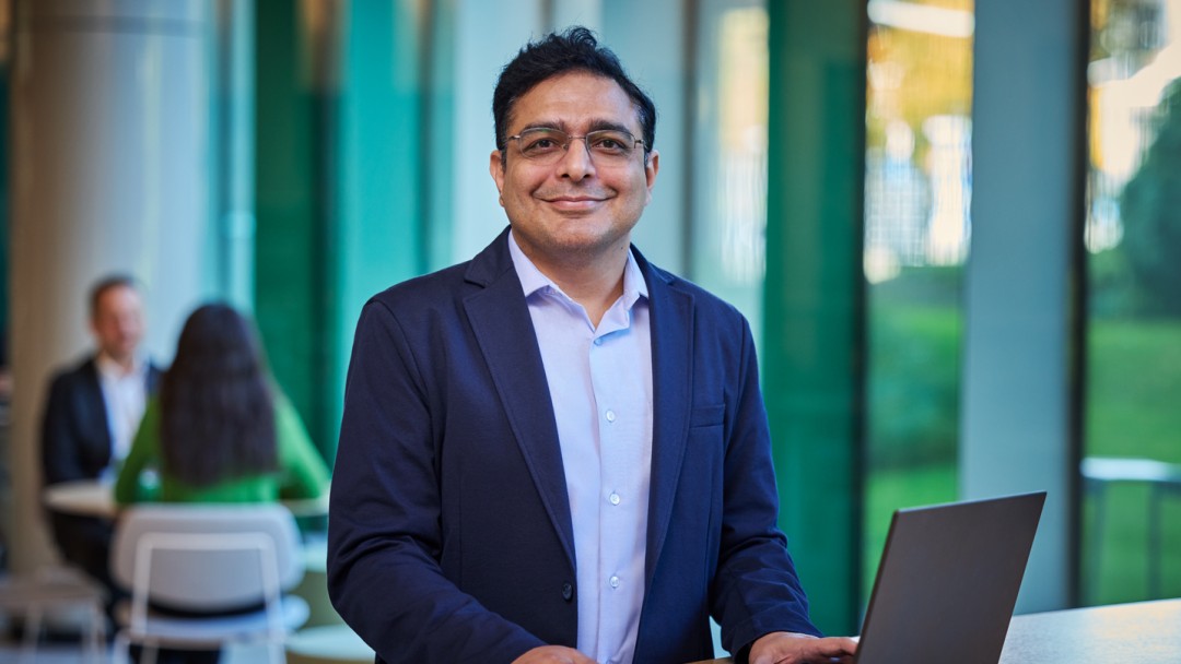 Ein Mitarbeiter der KfW Bankengruppe, Herr Dr. Raci Baghal, steht an einem Tisch mit einem aufgeklappten Laptop. Er trägt ein hellblaues Hemd, einen dukelblauen Sakko sowie eine Brille und schaut lächelnd in die Kamera. Im Hintergrund sitzen zwei Personen an einem Tisch. 