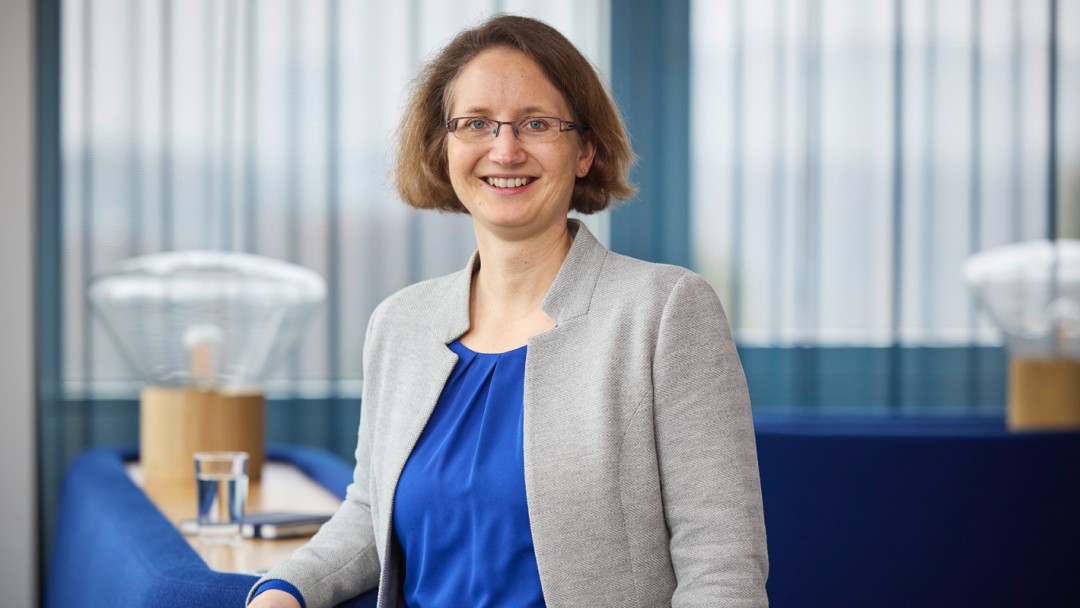 Elke Peetz, Mitarbeiterin der KfW Bankengruppe, steht angelehnt an einem Möbelstück. Sie trägt eine blaue Bluse, einen grauen Blzaer und eine Brille. Ihre kinnlangen Haare umrahmen ihr Gesicht. In der Hand hält sie einen zugeklappten schwarzen Laptop. Die Möbelstücke im Hintergrund sind zum Teil aus Holz und zum Teil in blau.
