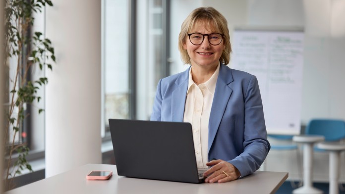 The KfW inclusion officer, Erika Holz, is standing in a bright room at a table on which an open laptop is placed. She has chin-length blonde hair, is wearing dark-rimmed glasses and a light blue blazer, and is looking friendly into the camera.