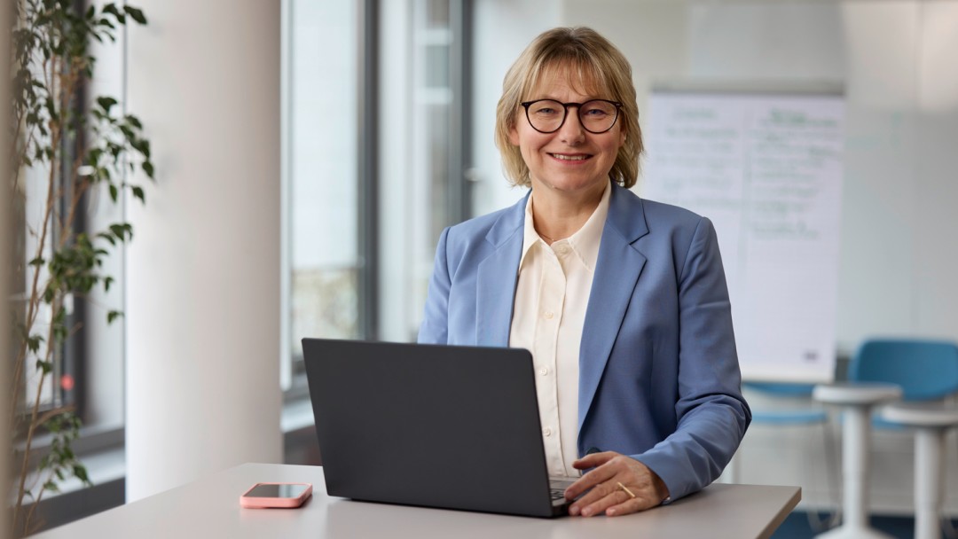 Die Inklusionsbeauftragte der KfW, Erika Holz, steht in einem hellen Raum an einem Tisch, auf welchem ein aufgeklappter Laptop steht. Sie hat kinnlange blonde Haare, trägt eine Brille mit dunklem Rahmen, einen hellblauen Blazer und schaut freundlich in die Kamera.