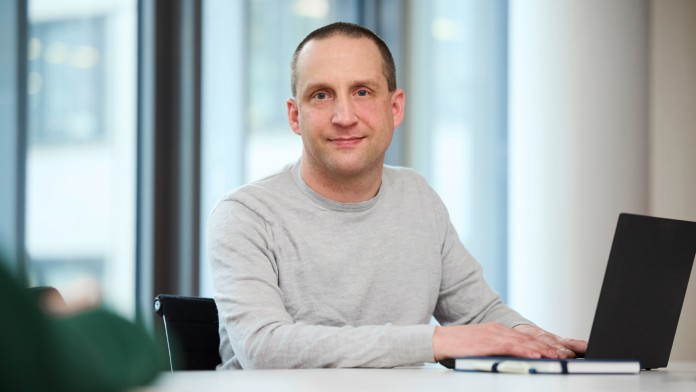 Gerald N., Mitarbeiter der KfW Bankengruppe, sitzt in einem grauen Pullover an eine Tisch. Er hat kurze braune Haare. Vor ihm steht ein aufgeklappter Laptop, daneben liegen ein Notizbuch und ein Kugelschreiber. Hinter ihm befindet sich eine große helle Fensterfront. 