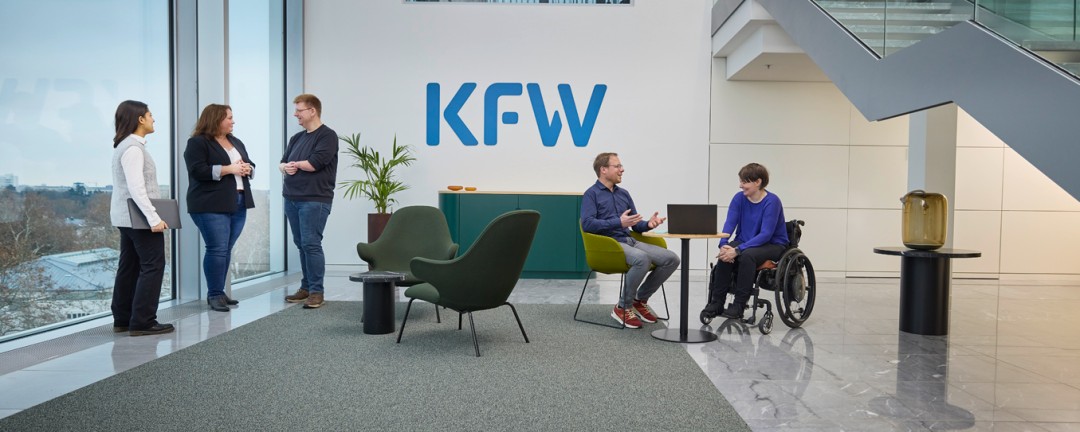 In einem hellen Raum mit einer großen Fensterfront ist auf der linken Seite an der hinteren Wand das blaue Logo der KfW zu sehen. Im Vordergrund befinden sich zwei Gruppen von Mitarbeitenden der KfW. Auf der rechten Seite sitzt ein Mann auf einem Stuhl und eine Frau im Rollstuhl an einem Tisch mit einem aufgeklappten Laptop. Auf der linken Seite steht eine Gruppe von drei Personen, die sich unterhalten. Zur Gruppe gehören zwei Frauen mit dunklen Haaren und ein Mann mit fehlender linker Hand. 