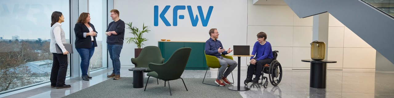 In einem hellen Raum mit einer großen Fensterfront ist auf der linken Seite an der hinteren Wand das blaue Logo der KfW zu sehen. Im Vordergrund befinden sich zwei Gruppen von Mitarbeitenden der KfW. Auf der rechten Seite sitzt ein Mann auf einem Stuhl und eine Frau im Rollstuhl an einem Tisch mit einem aufgeklappten Laptop. Auf der linken Seite steht eine Gruppe von drei Personen, die sich unterhalten. Zur Gruppe gehören zwei Frauen mit dunklen Haaren und ein Mann mit fehlender linker Hand. 